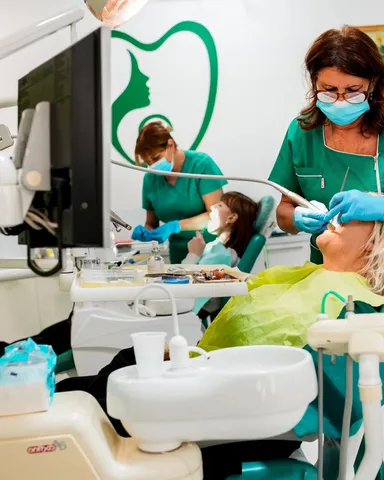 Doktorka sa naočarima leči pacijenta, iza nje koleginica sa detetom; na zidu zeleni logo ordinacije